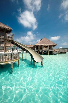 a water slide in the middle of some clear blue water with huts on stilts