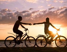 two people riding bikes on the beach at sunset