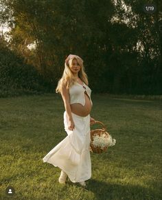 a pregnant woman in a white dress is holding a basket and walking through the grass