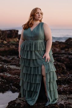 a woman in a green dress standing on rocks near the ocean at sunset with her legs spread out