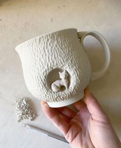 a hand holding a white ceramic cup with a small animal on it's side