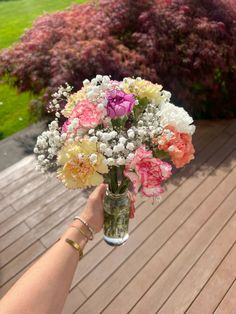 a person holding a vase filled with flowers on top of a wooden deck next to bushes