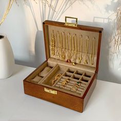 an open wooden box filled with gold jewelry on top of a white table next to a vase