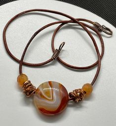 This vibrant necklace features a striking Sardonyx Agate pendant, known for its bold bands of color, complemented by warm copper and rich Carnelian beads. Strung on a durable leather cord, the natural textures and tones create a perfect balance of earthy elegance and bright, energetic design. Ideal for those who love statement pieces with a touch of artisanal flair, this necklace adds a pop of color and warmth to any outfit. Approximately 20" long. For reference Sardonyx Agate in almost 1" aroun Adjustable Carnelian Round Pendant Necklace, Adjustable Carnelian Necklace Hand Wrapped, Adjustable Hand Wrapped Carnelian Necklace, Artisan Brown Necklace With Waxed Cord, Brown Agate Hand Wrapped Necklace, Adjustable Wire Wrapped Carnelian Necklace, Earthy Elegance, Love S, Textures And Tones