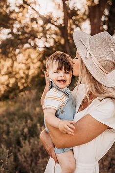 a woman holding a child in her arms and kissing him on the cheek with trees in the background