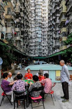 several people sitting around a table in an open area with tall buildings behind them and one person standing