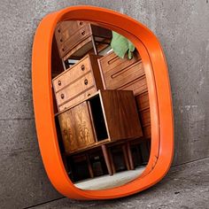 an orange mirror reflecting several pieces of furniture on the floor in front of a concrete wall