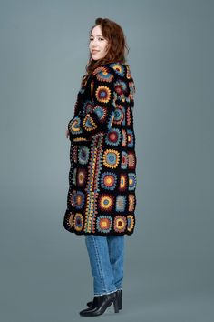 a woman standing in front of a gray background wearing a black and multicolored crocheted coat