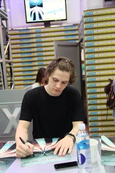 a young man sitting at a table in front of a computer monitor and writing on paper