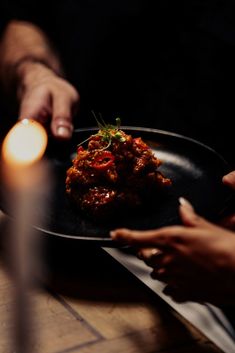 two hands reaching for food on a black plate with candles in the background and one hand holding a lit candle