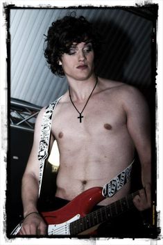 a shirtless young man holding a red guitar in his left hand and wearing a cross necklace