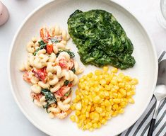 a white plate topped with pasta, corn and spinach next to an egg on a table