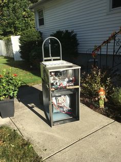 a metal box sitting on the side of a sidewalk next to flowers and plants in front of a house