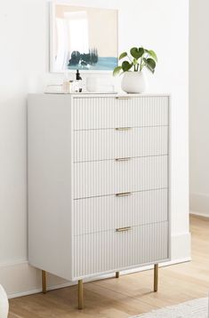 a white chest of drawers sitting next to a plant on top of a wooden floor