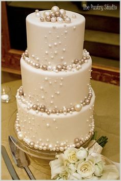 a three tiered wedding cake with pearls and flowers on the table next to it