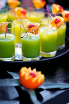 small glasses filled with green liquid and fruit garnished with toothpicks on a black tray