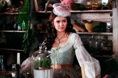 a woman dressed in period clothing sitting at a table with plants and bottles on it