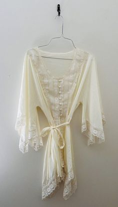 Indulge in vintage charm with this stunning 70s Val Mode lingerie short robe / nightgown . The satiny cream fabric is beautifully accented with delicate lace trim, a tie waist, and button front adding a touch of sleek sophistication to your sleepwear routine. This original piece is perfect for anyone looking to add a unique touch to their wardrobe. Can be worn in the bedroom or out! Crafted with high-quality nylon material, this robe is both comfortable and durable. The ivory color and regular size type make it a versatile addition to any vintage clothing collection. Made in the United States, this original piece is a must-have for any fashion-forward individual. Sized as a medium but does run small. Fits more like a small. Measurements: Length 30" Shoulder to shoulder 15" Waist 14" Feminine Lace Robe For Sleep, Cream Lace-trim Sleepwear For Wedding, Beige Lace Trim Sleepwear For Summer, Summer Beige Lace Trim Sleepwear, Cream Wedding Sleepwear With Lace Trim, White Vintage Sleepwear With Delicate Lace, Feminine Cream Nightgown With Lace Trim, White Bohemian Nightgown For Loungewear, Cottagecore White Nightgown With Lace Trim