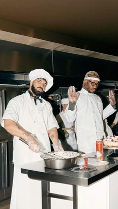 two men in chef's uniforms are preparing food on a counter top, while one man holds his hand up