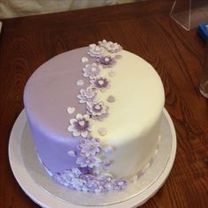 a white and purple cake with flowers on the top is sitting on a wooden table