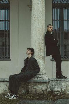 two young men sitting on the side of a pillar in front of a white building