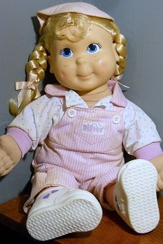 a doll with blonde hair and blue eyes sitting on a wooden table next to a gray wall