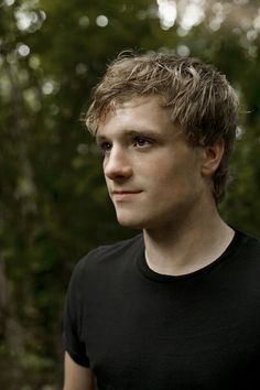 a young man in a black shirt is looking into the distance with trees behind him