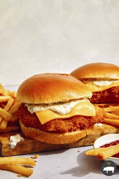 two chicken sandwich sandwiches with cheese and french fries on a cutting board next to some ketchup