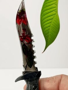 a hand holding a knife with red flowers in it and a green leaf sticking out of the blade