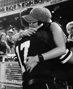 two people hugging each other in front of a crowd at a sporting event, one is wearing a baseball cap and the other has a cell phone