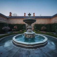 a fountain in the middle of a courtyard surrounded by hedges and bushes with pink flowers on either side