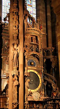 an ornate clock in the middle of a building with stained glass windows behind it and statues on either side