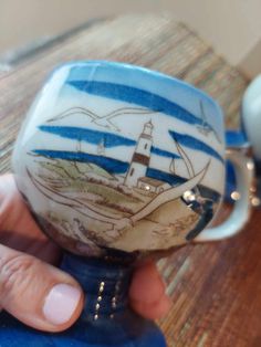 a hand holding a blue and white coffee cup with a lighthouse on the side, in front of a wooden table