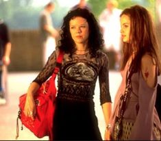 two women standing next to each other on a sidewalk with people in the background and one woman carrying a red purse