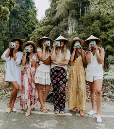 four women are taking pictures with their cell phones while wearing hats and dresses in front of some trees