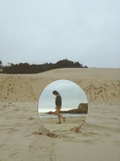 a person walking on the beach in front of a round mirror that is reflecting their reflection