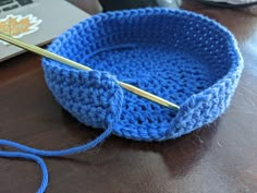 a blue crocheted bowl with a knitting needle in it on a table next to a laptop