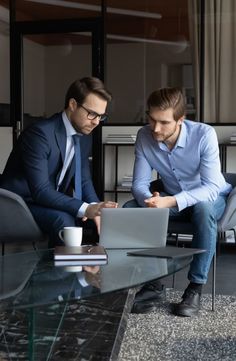 two men in suits sitting at a glass table looking at a laptop computer, both on their knees