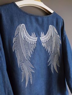 the back of a denim shirt with white embroidered wings on it, hanging from a hanger