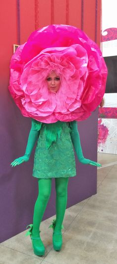 a woman dressed in green and pink holding a large paper flower over her head while standing next to a purple wall