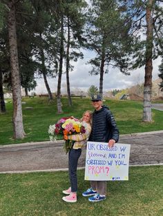 a man and woman standing next to each other in front of trees holding a sign