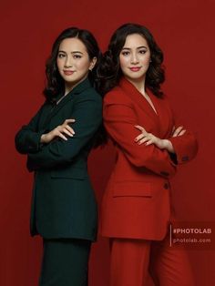 two women standing next to each other in front of a red wall with their arms crossed