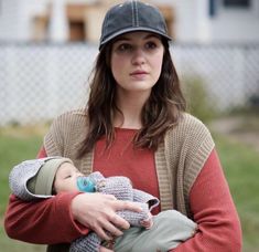 a woman holding a baby wearing a hat and red shirt in front of a house