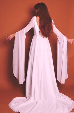 a woman wearing a white dress with long sleeves and an open back, standing in front of a brown background