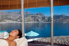 a woman laying in a bathtub looking out at the ocean and mountains from a hotel room