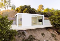 a small white house sitting on top of a hill next to some trees and bushes