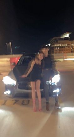 two people standing in front of a car at night with their arms around each other