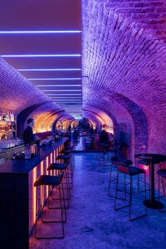 an underground bar with purple lighting and stools in the foreground, while people are seated at tables on either side