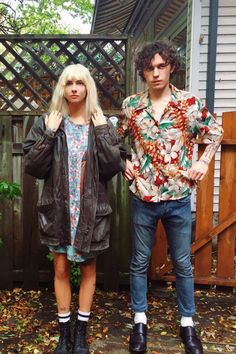 two people standing next to each other in front of a fence with leaves on the ground