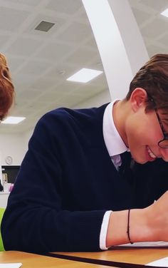 two people sitting at a table with papers and pens in front of them, one man is smiling